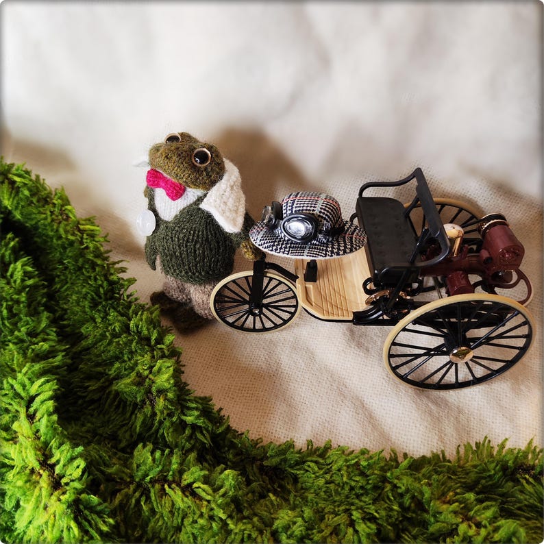 May include: A whimsical scene featuring a knitted frog wearing a jacket, bow tie, and a white collar, standing next to a miniature vintage car. The car has a black seat, wooden body, and black wheels. A green, textured border frames the scene.