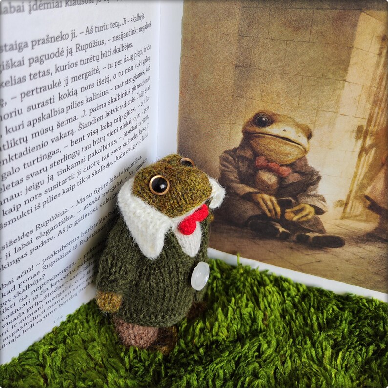 May include: A handmade, knitted frog figurine wearing a green coat, white collar, and red bow tie. The frog has large, round eyes and is positioned on a bed of green moss, next to an open book with text and an illustration of a frog.