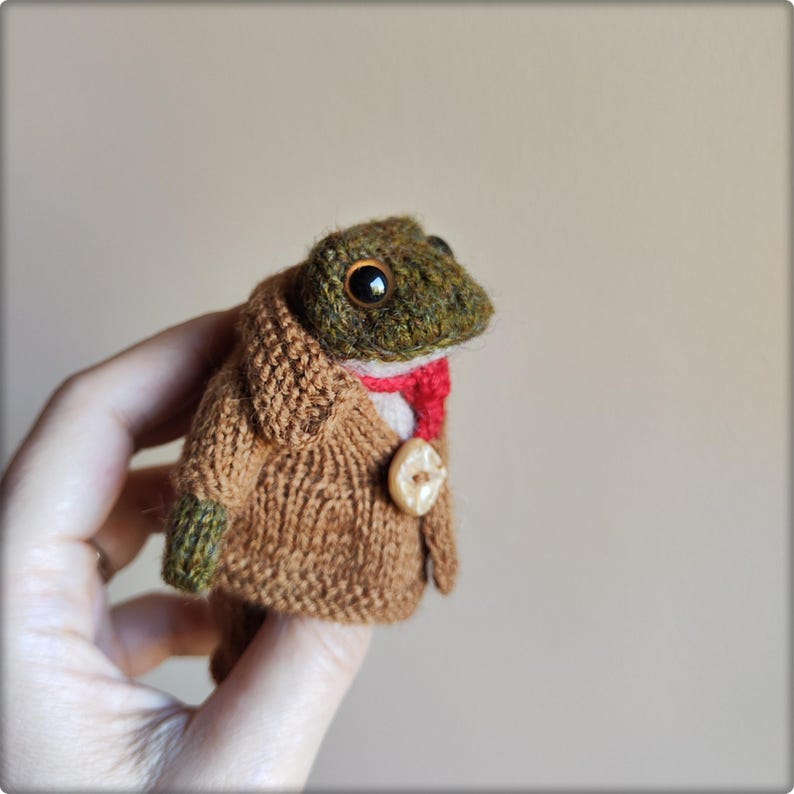 May include: A small, handcrafted knitted frog figurine featuring a textured olive green head with large dark eyes. The frog wears a tan knitted coat with a single light-colored button, a bright red necktie, and matching olive green knitted sleeves.