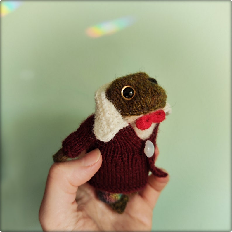 May include: A handmade, knitted frog doll wearing a burgundy sweater, white collar, and a red bow tie. The frog has large, dark eyes and a button closure. The frog's head is brown and green.