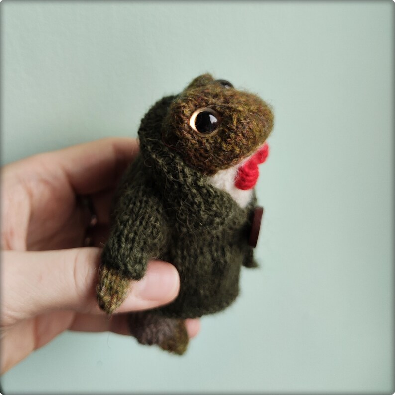 May include: A handmade, knitted frog doll wearing a green jacket with a red bow tie and a white shirt collar. The frog has large, expressive eyes and a button closure on the jacket. The doll is a unique, handcrafted item.