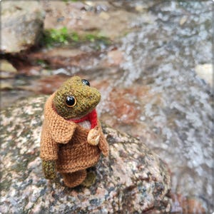May include: A small, handcrafted crochet frog figurine with a textured olive green body and large, glossy black eyes. The frog wears a miniature, tailored brown knit coat with a wide collar, a thin red scarf, and a small, light-colored wooden button.
