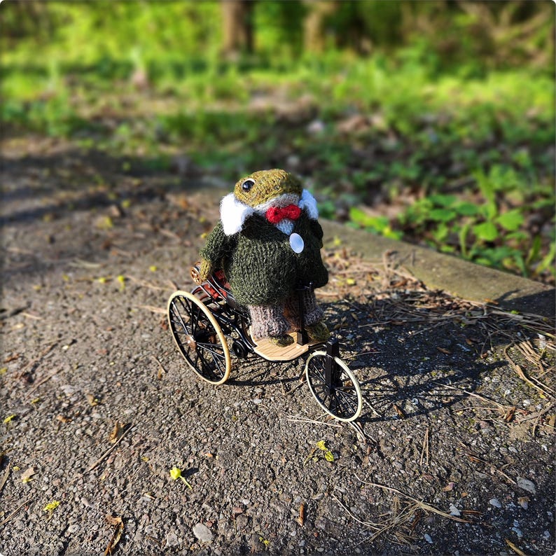 May include: A handmade knitted frog wearing a green jacket, white collar, and red bow tie, riding a vintage-style tricycle. The frog has a green and beige hat. The tricycle has black wheels and a wooden seat.