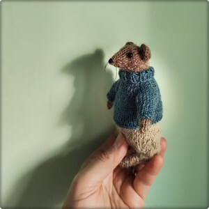 May include: A small, hand-knitted mouse figurine featuring brown yarn fur, black bead eyes, and thin whiskers. The mouse is wearing a textured blue knitted sweater with a high collar and light beige knitted trousers, crafted with visible yarn stitches.
