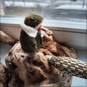 May include: A handmade felt frog figurine in shades of green, brown, and white, perched on a piece of driftwood. The frog has large, round eyes, a white scarf, and a red accent. A woven object is in the foreground.