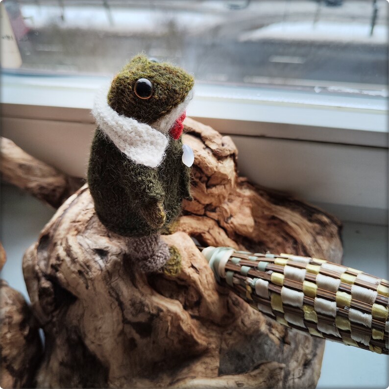 May include: A handmade felt frog figurine in shades of green, brown, and white, perched on a piece of driftwood. The frog has large, round eyes, a white scarf, and a red accent. A woven object is in the foreground.