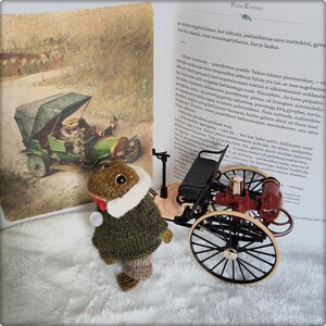 May include: A handmade crochet frog wearing a green sweater, white collar, and brown pants. A small toy antique car is next to the frog. A book with an illustration of a car is in the background.