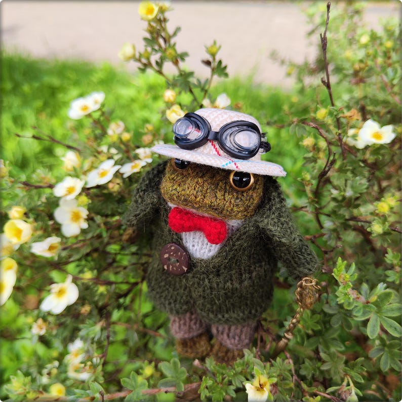 May include: A handcrafted, knitted frog doll dressed in a green jacket, red bow tie, and a white hat with goggles. The frog is holding a small, ornate cane and is surrounded by white flowers and green foliage.