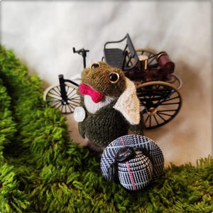 May include: A handcrafted frog figurine wearing a suit, bow tie, and a hat with goggles. The frog is positioned in front of a miniature bicycle and a vintage car model, set against a backdrop of green moss.