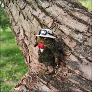 May include: A handmade, whimsical frog doll wearing a green jacket, pants, and a red bow tie. The frog is accessorized with a white hat with goggles and a pocket watch chain. The doll is posed against a tree trunk.