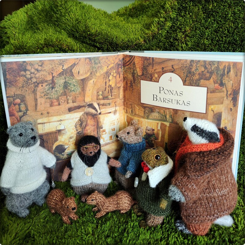 May include: A collection of handmade knitted animal figures, including a badger, otter, and mouse, are arranged in front of an open book. The book's page features an illustration with the text "Ponas Barsukas". The figures are on a green surface.