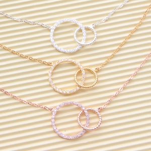 Peut inclure: Trois colliers délicats avec des pendentifs en forme de cercles entrelacés. Chaque pendentif est orné de pierres de zircone cubique claires scintillantes. Les colliers sont disponibles en argent, or et or rose.