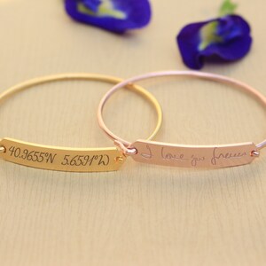May include: Two gold bangle bracelets with engraved bars. One bar reads "40.9655°N 5.6591°W" and the other bar reads "I love you forever".