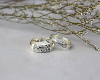 May include: Two silver wedding bands with fingerprint engravings. The band on the left has the inscription "Mark & Jan" on the inside.