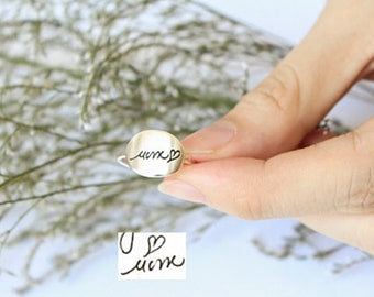 Puede incluir: Un anillo de plata con una parte superior redonda y plana. El anillo presenta la palabra manuscrita "mom" con un símbolo de corazón. También es visible una segunda nota manuscrita con las palabras "U mom". El anillo se sostiene sobre un fondo de vegetación.