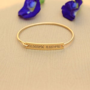 Puede incluir: Un brazalete de oro con una pequeña etiqueta rectangular grabada con las coordenadas "51.5072°N 0.1274°W".
