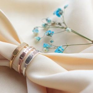 Peut inclure: Trois bracelets en or, argent et or rose avec une empreinte digitale, dont l'un porte l'inscription "Love & Grace". Les bracelets sont sur un fond de tissu beige clair.