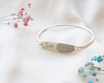 May include: Silver bangle bracelet with a personalised oval charm engraved with the name "Bells" and a fingerprint.