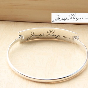 Könnte beinhalten: Ein silbernes Armreif-Armband mit einer personalisierten Inschrift einer Unterschrift, die "Janet Hayne" lautet.