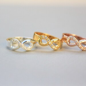 May include: Three infinity symbol rings with small clear stones set in the metal. The rings are in silver, gold, and rose gold.
