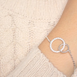 Op de afbeelding: Een zilveren armband met twee in elkaar grijpende cirkels, waarvan er één is bezet met heldere zirkonia-stenen. De armband wordt om de pols gedragen.