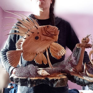 K&ouml;nnte beinhalten: Eine Holzskulptur eines Fisches mit einer stacheligen Flosse, einem Anker und einem Netz auf einem Holzsockel. Die Skulptur ist in Braun- und Beiget&ouml;nen bemalt.