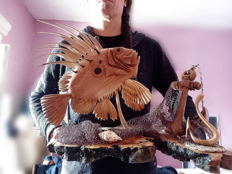 K&ouml;nnte beinhalten: Eine Holzskulptur eines Fisches mit einer stacheligen Flosse, einem Anker und einem Netz auf einem Holzsockel. Die Skulptur ist in Braun- und Beiget&ouml;nen bemalt.