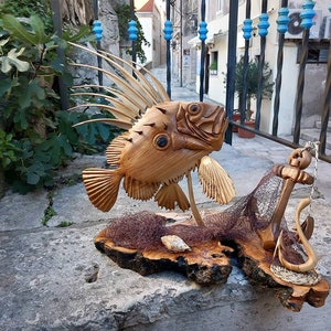 K&ouml;nnte beinhalten: Eine Holzskulptur eines Fisches mit einer stacheligen Flosse, einem Fischernetz und einem Anker. Die Skulptur ist aus hellem braunem Holz gefertigt und auf einem Holzsockel ausgestellt.
