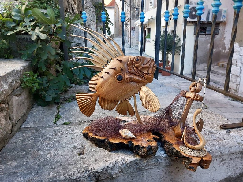 K&ouml;nnte beinhalten: Eine Holzskulptur eines Fisches mit einer stacheligen Flosse, einem Fischernetz und einem Anker. Die Skulptur ist aus hellem braunem Holz gefertigt und auf einem Holzsockel ausgestellt.