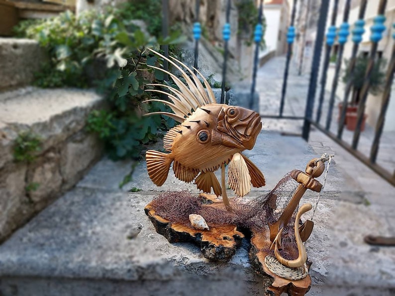 K&ouml;nnte beinhalten: Eine Holzskulptur eines Fisches mit einer stacheligen Flosse, einem Anker und einem Netz auf einem Holzsockel.