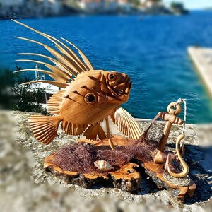 K&ouml;nnte beinhalten: Eine Holzskulptur eines Fisches mit einer stacheligen Flosse, einem Anker und einem Fischernetz auf einem Holzsockel. Die Skulptur ist vor dem Hintergrund eines blauen Ozeans und eines Steinpiers platziert.