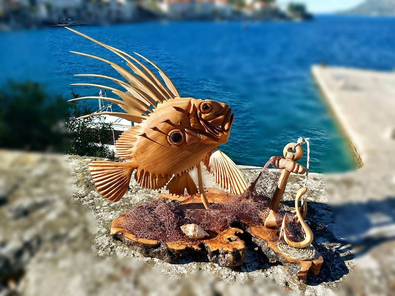 K&ouml;nnte beinhalten: Eine Holzskulptur eines Fisches mit einer stacheligen Flosse, einem Anker und einem Fischernetz auf einem Holzsockel. Die Skulptur ist vor dem Hintergrund eines blauen Ozeans und eines Steinpiers platziert.
