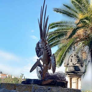 K&ouml;nnte beinhalten: Eine detaillierte Holzskulptur eines Fisches mit einer stacheligen Flosse, die auf einem Steinsockel ausgestellt ist. Die Skulptur steht vor einem Palmenbaum und einem Steinbau mit einem Wappen.