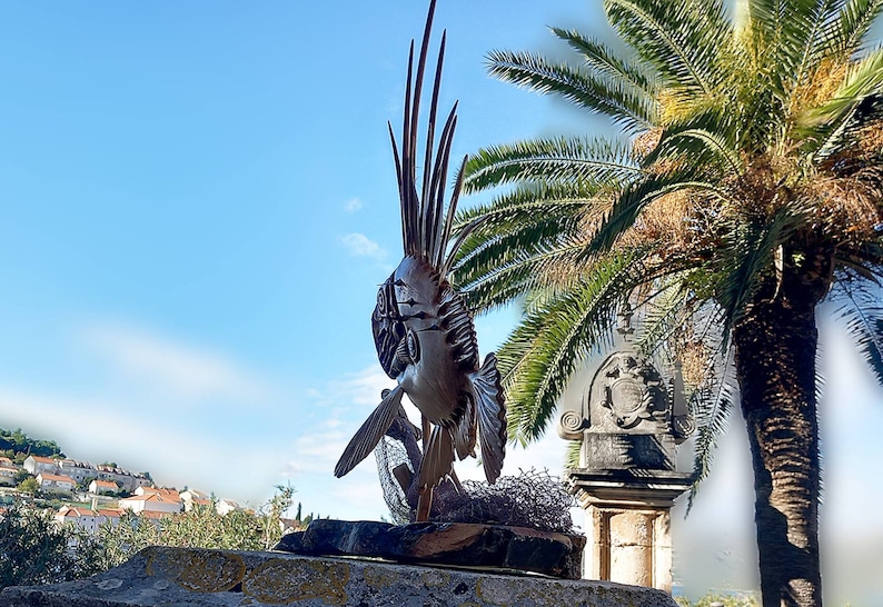 K&ouml;nnte beinhalten: Eine detaillierte Holzskulptur eines Fisches mit einer stacheligen Flosse, die auf einem Steinsockel ausgestellt ist. Die Skulptur steht vor einem Palmenbaum und einem Steinbau mit einem Wappen.