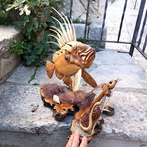 K&ouml;nnte beinhalten: Eine Holzskulptur eines Fisches mit einer stacheligen Flosse, die auf einem Holzsockel mit einem Fischernetz ruht. Die Skulptur ist in Braun- und Beiget&ouml;nen bemalt.