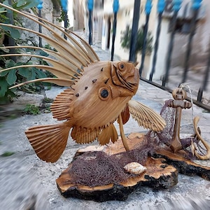 K&ouml;nnte beinhalten: Eine Holzskulptur eines Fisches mit einer stacheligen Flosse, einem Anker und einem Fischernetz auf einem Holzsockel.