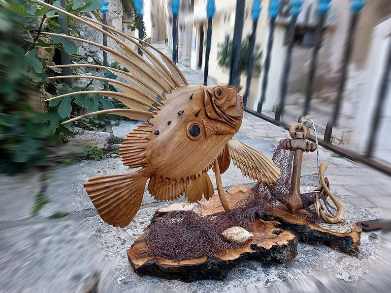 K&ouml;nnte beinhalten: Eine Holzskulptur eines Fisches mit einer stacheligen Flosse, einem Anker und einem Fischernetz auf einem Holzsockel.