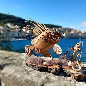 K&ouml;nnte beinhalten: Eine Holzskulptur eines Fisches mit einer langen, stacheligen Flosse, einem Netz und einem Anker. Die Skulptur steht auf einem Holzsockel und ist vor einem verschwommenen Hintergrund einer K&uuml;stenstadt platziert.