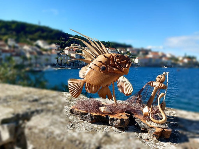 K&ouml;nnte beinhalten: Eine Holzskulptur eines Fisches mit einer langen, stacheligen Flosse, einem Netz und einem Anker. Die Skulptur steht auf einem Holzsockel und ist vor einem verschwommenen Hintergrund einer K&uuml;stenstadt platziert.