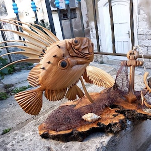 K&ouml;nnte beinhalten: Eine Holzskulptur eines Fisches mit einer langen, stacheligen R&uuml;ckenflosse, einem Anker und einem Fischernetz auf einem Holzsockel.