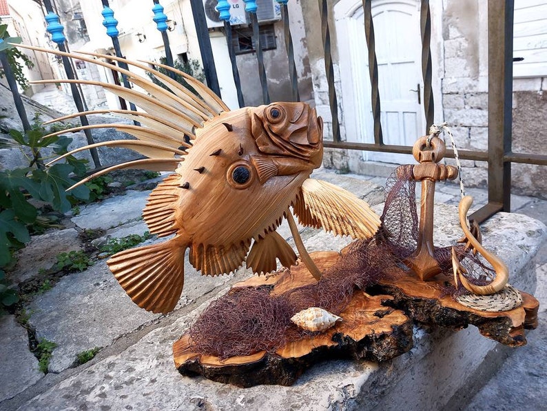K&ouml;nnte beinhalten: Eine Holzskulptur eines Fisches mit einer langen, stacheligen R&uuml;ckenflosse, einem Anker und einem Fischernetz auf einem Holzsockel.