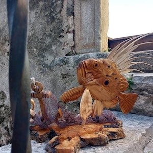K&ouml;nnte beinhalten: Eine Holzskulptur eines Fisches mit einer langen, stacheligen R&uuml;ckenflosse. Der Fisch ist auf einem Holzsockel mit einem St&uuml;ck Netz montiert.
