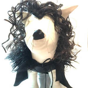 May include: A handmade plush doll with a white face, ears, and legs. The doll has black curly hair, a black nose, and a black cape tied with a black string. The doll is a unique, handcrafted item.