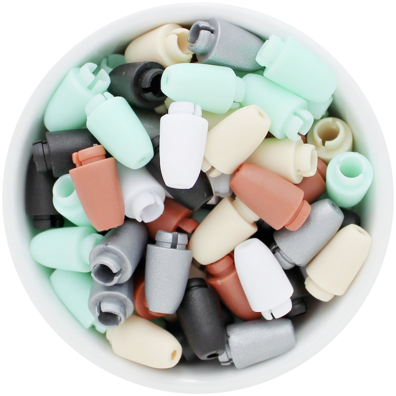 May include: A white bowl filled with a variety of colorful, small, plastic objects. The objects are in shades of white, black, gray, brown, and mint green. The objects have a unique shape with a cylindrical body and a notched top.