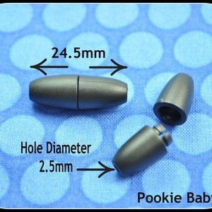 May include: Dark gray magnetic clasp with a 24.5mm length and a 2.5mm hole diameter. The clasp is shown in three pieces, with one fully assembled and two disassembled. The background is a blue fabric with white polka dots. The brand name "Pookie Baby" is visible.