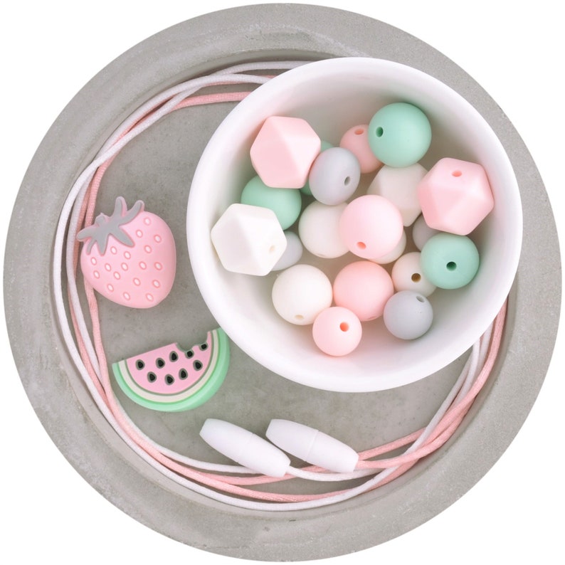 May include: A set of silicone beads in pink, green, and gray colors, with a strawberry and watermelon design, in a white bowl on a gray tray. The beads are strung on a white and pink cord.