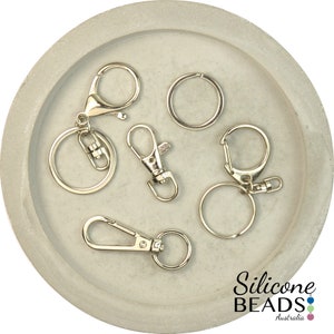 May include: A collection of six silver key rings and clasps, including a split ring, a lobster clasp, and a swivel clasp. The key rings are displayed on a gray concrete surface.