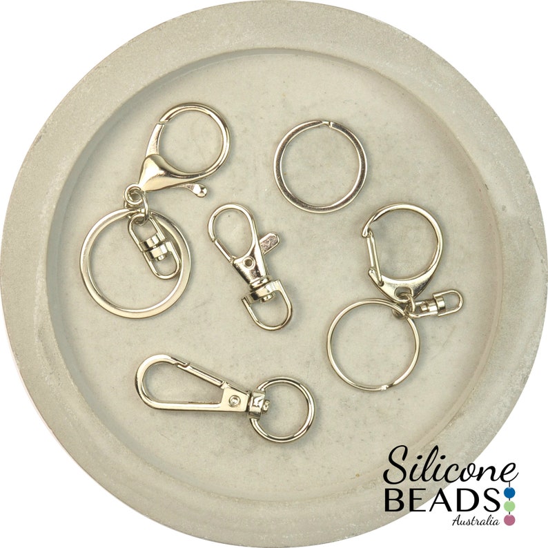 May include: A collection of six silver key rings and clasps, including a split ring, a lobster clasp, and a swivel clasp. The key rings are displayed on a gray concrete surface.