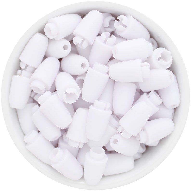 May include: A close-up shot of a white bowl filled with numerous white plastic cord locks. The cord locks are cylindrical with a rounded top and a hole for the cord. Some have a slotted design.