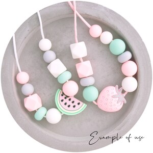 May include: Three silicone teething necklaces with different fruit charms. The necklaces are pink, gray, and white with green accents. The charms are a watermelon, a strawberry, and a square.